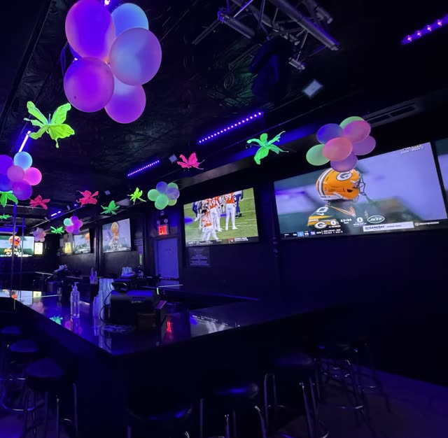 The long bar at Sweets of NY: stools along the counter, pink and purple balloons and neon butterflies overhead, and football on a wall of big screens.