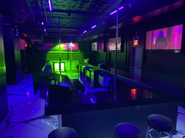 The stage end of the room under black light: the pole, the padded wall behind it lit green, screens along the walls and the marble floor.